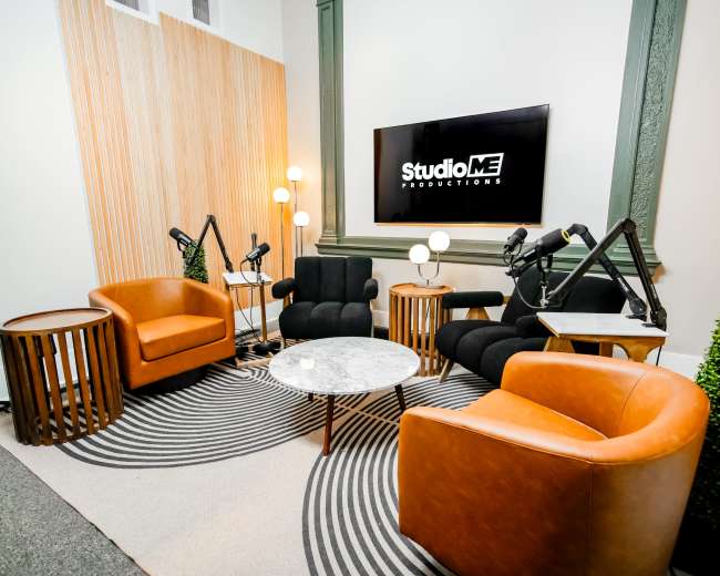 The image shows a modern studio setting with two brown armchairs, black upholstered chairs, a marble coffee table, and a circular rug, all arranged around a large screen displaying the "StudioME Productions" logo.