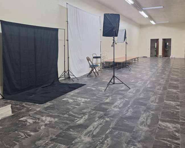 The image shows a spacious room with a light-colored floor, equipped with photography backdrops, lighting equipment, and a long table.
