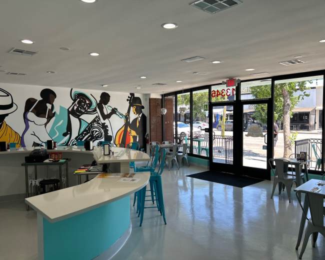 The image shows a modern cafe interior with colorful wall murals, white countertops, and metal seating arranged around tables.