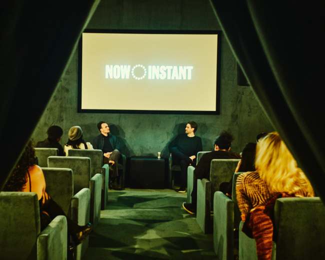 A small audience is seated in a theater, watching two speakers on stage with a screen displaying "NOW INSTANT" behind them.