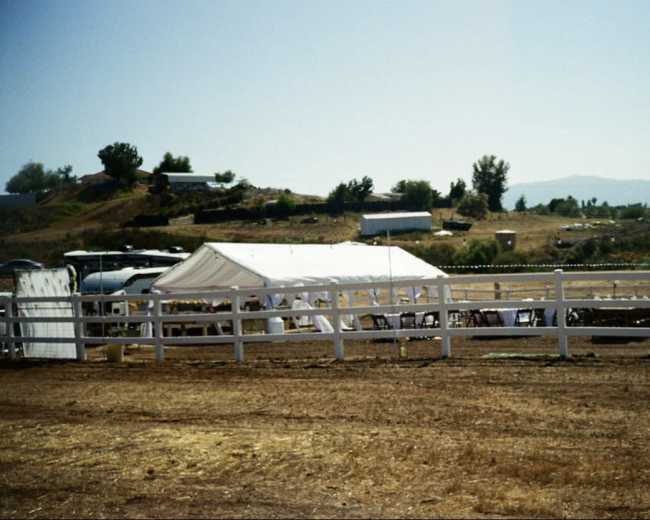 A white tent is set up in an open field with a fenced area and several buildings on a hillside in the background.