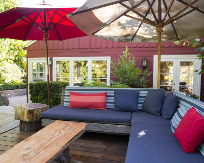 A backyard seating area features striped sofas, red umbrellas, and a wooden table.