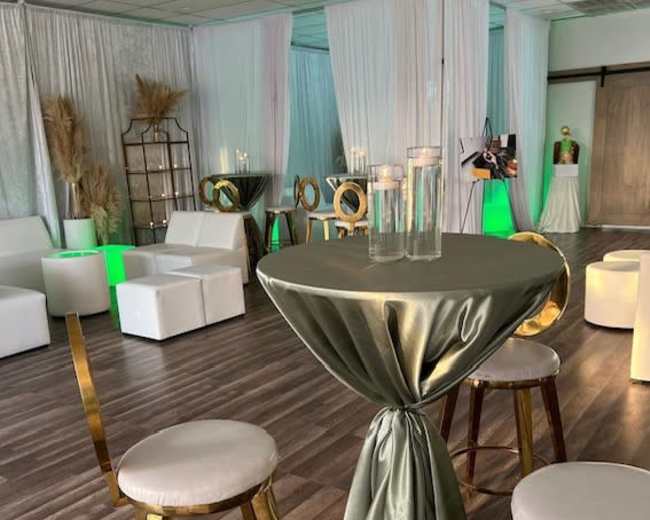 A decorated event space with white drapes, modern seating, and a table draped in a greenish cloth with candles.