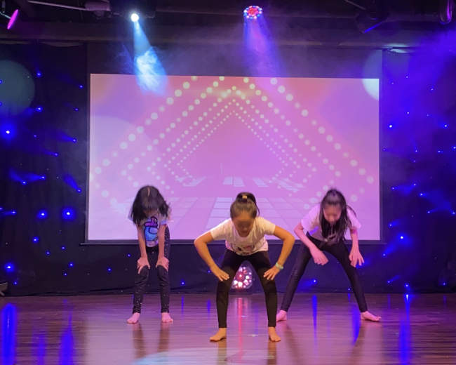 Three children perform a dance routine on stage with a colorful backdrop and stage lights.