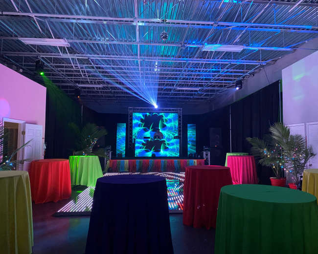 A colorful party space features several round tables with tablecloths in red, green, and yellow, illuminated by a backdrop of vibrant lights and a projector display.