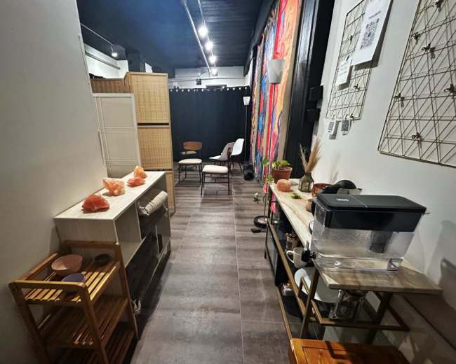 The image shows a narrow, well-decorated room with wooden carts, a water dispenser, and colorful wall hangings, leading to a seating area at the back.