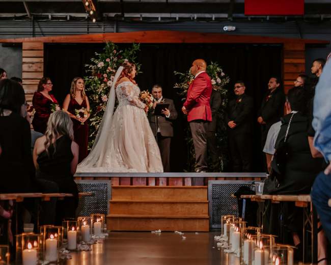 A bride and groom stand at the altar during their wedding ceremony, surrounded by their wedding party and decorated with floral arrangements and candles lining the aisle.