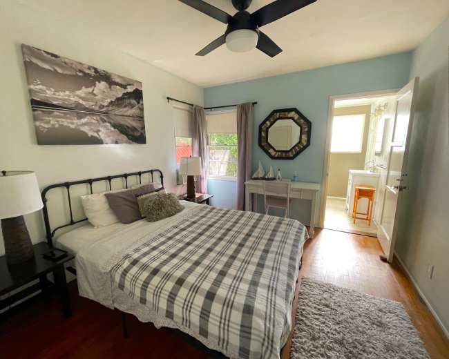 The image shows a well-lit bedroom featuring a bed with a checkered blanket, a nightstand with a lamp, a vanity area, and an en-suite bathroom visible in the background.