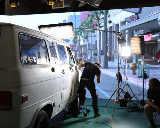 A person is adjusting equipment on the side of a vintage white van in a studio setting with lighting and a city backdrop.