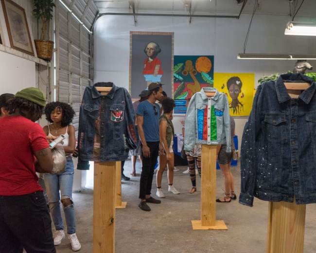 The image shows a bustling art gallery with people interacting while denim jackets displayed on wooden stands are featured prominently in the foreground.