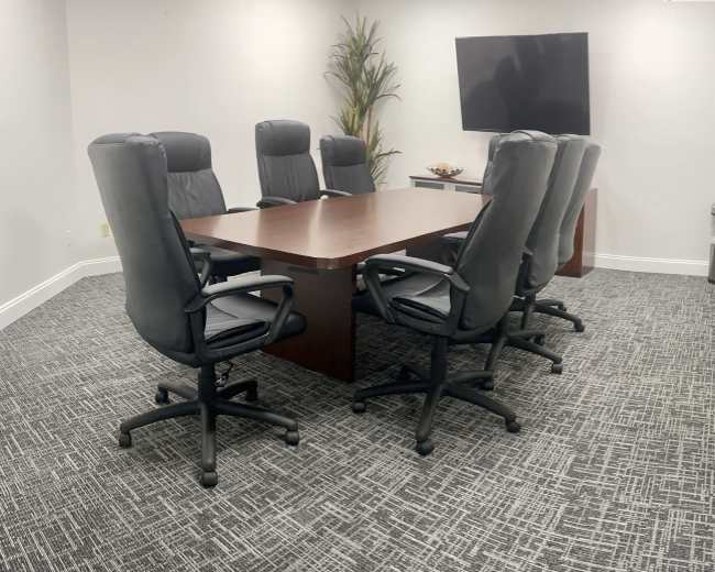 A conference room features a large rectangular table surrounded by six ergonomic chairs, with a television mounted on the wall and a potted plant in the corner.
