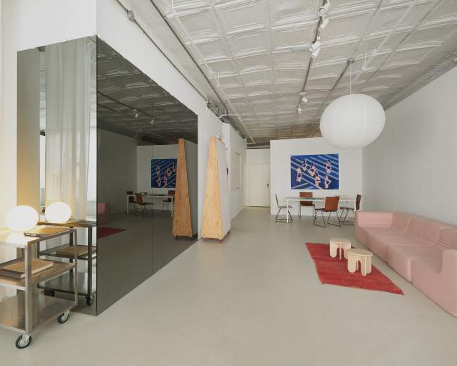 The image depicts a modern, minimalist interior space featuring a pink sofa, a mirrored wall, wooden dividers, and a dining area with a round paper lantern hanging from the ceiling.