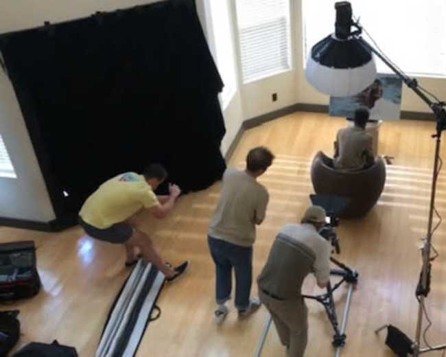 A photography crew sets up a shoot in a well-lit room featuring a model seated in a large round chair, with various equipment positioned around them.