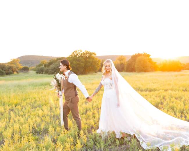 A couple holds hands in a field during sunset, with the bride wearing a long veil and the groom in a suit.