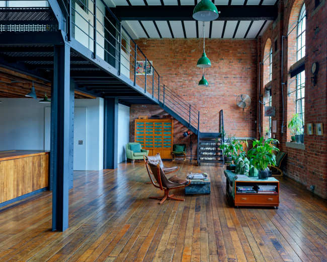 The image shows a spacious industrial-style loft featuring exposed brick walls, large windows, wooden floors, and a mix of modern and vintage furniture.