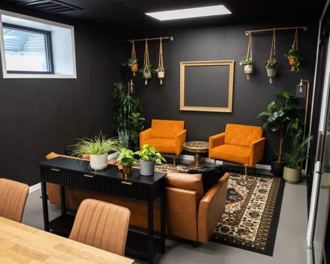 The image features a modern office space with two orange armchairs, a dark wooden table, plants, and a decorative rug.