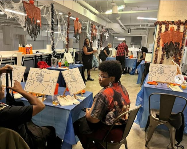 Two individuals sit at a table adorned with canvases and art supplies in a decorated room featuring Halloween-themed decorations and people engaged in painting activities.