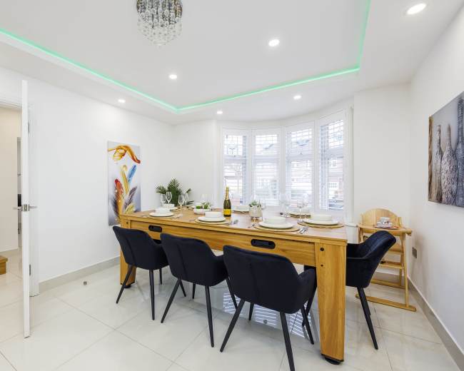 A modern dining room features a long wooden table set with dinnerware and surrounded by black chairs, with large windows and decorative artwork on the walls.