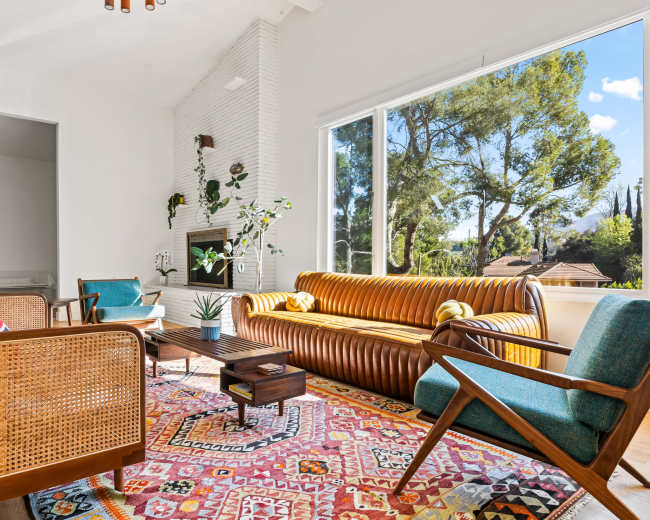 The brightly lit living room features a modern design with a leather couch, two teal armchairs, a wooden coffee table, and a colorful area rug, all overlooking a view of trees through large windows.