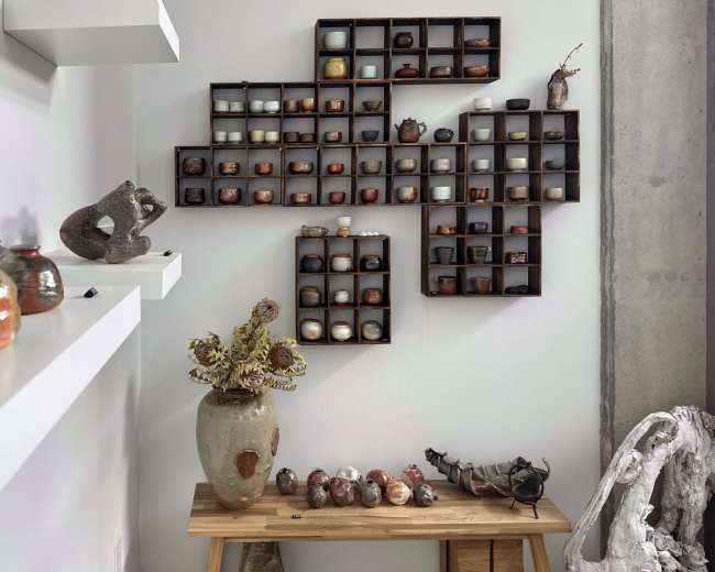 The image shows a wall display featuring various ceramic items arranged in boxes, along with a wooden table that holds additional pottery and sculptures.