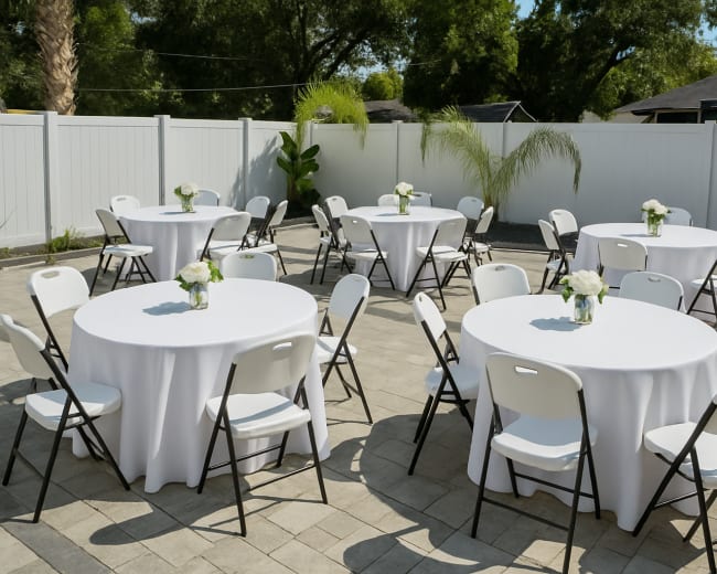 The image shows a well-arranged outdoor patio with several round tables, each covered with white tablecloths and featuring a small floral centerpiece.