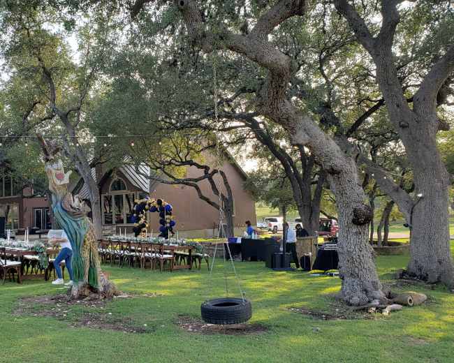 A large outdoor event setup features a long table with chairs beneath trees, adjacent to a building and decorated areas with balloons.