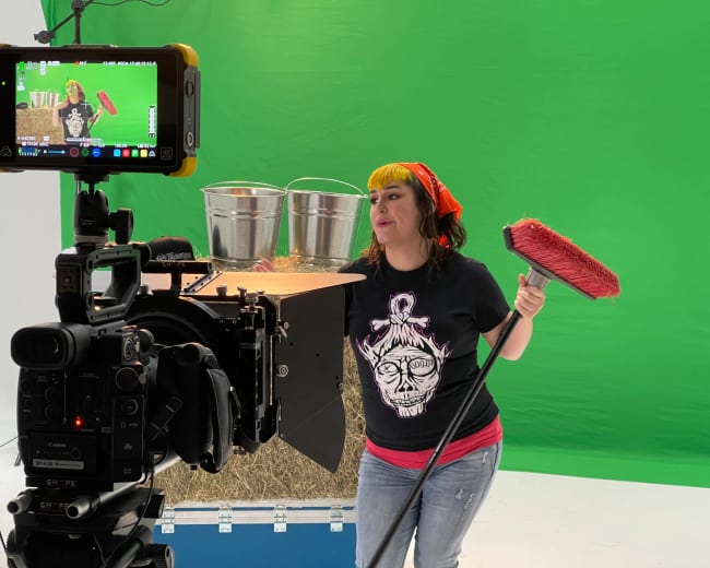 A woman in a black shirt and red bandana stands in front of a green screen, holding a broom and posing near buckets on a set with hay.