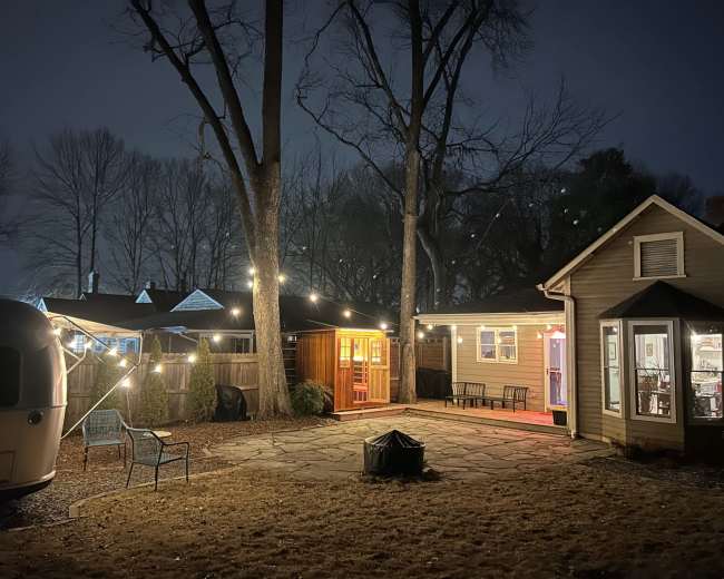 The image shows a backyard at night with string lights illuminating a stone patio area, a trailer on one side, and a house with warm light coming from the windows.