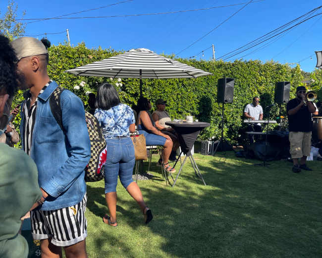 A gathering on a green lawn features people socializing under umbrellas while a band plays music in the background.