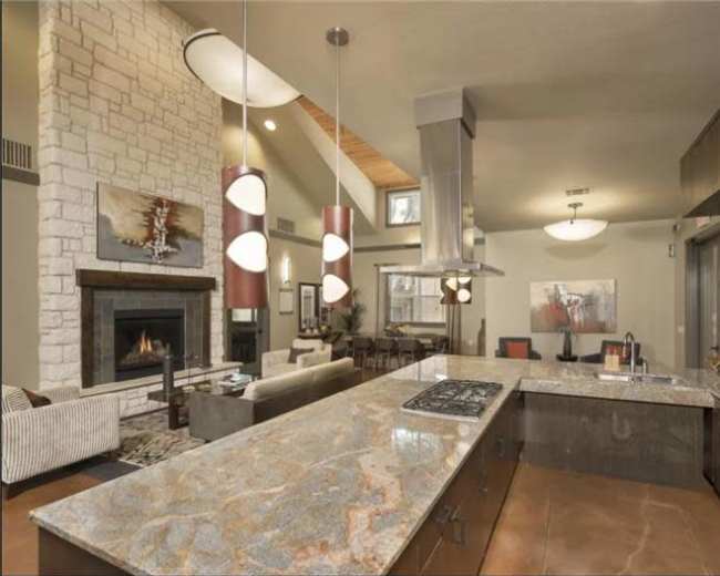 The spacious kitchen features a large granite island with a stovetop, modern fixtures, and a view of a cozy living area with a fireplace.
