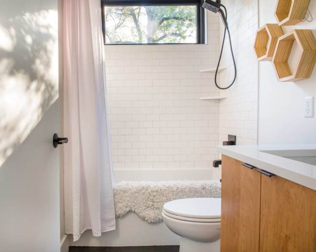 A bright bathroom features white tile walls, a shower with a glass window, and a wooden cabinet below a white countertop.