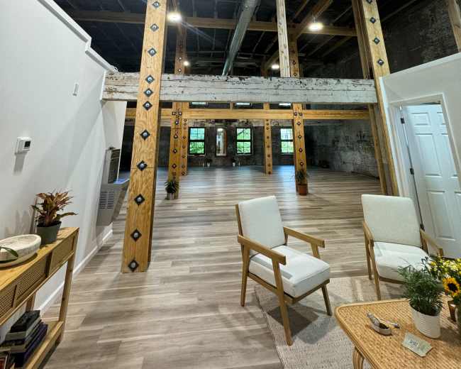 An interior space with wooden support beams, large windows, and minimal furniture, featuring a light-colored floor and exposed industrial elements.