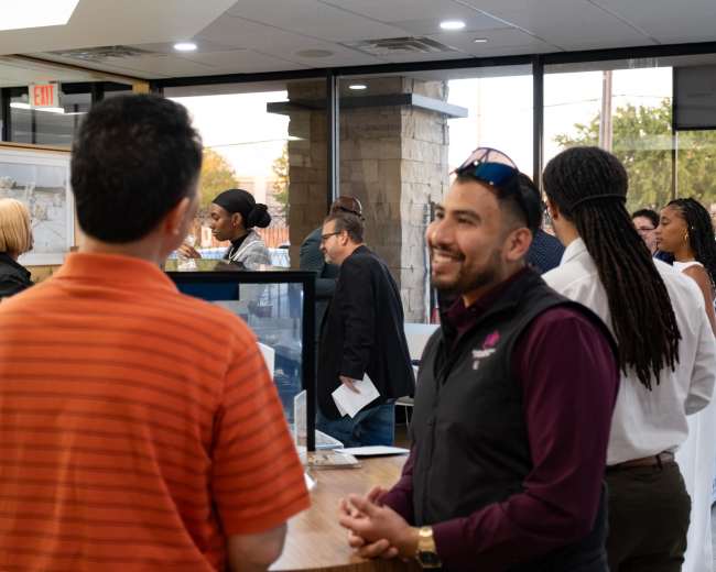 A group of people is engaged in conversation in a modern office setting with a reception area visible in the background.