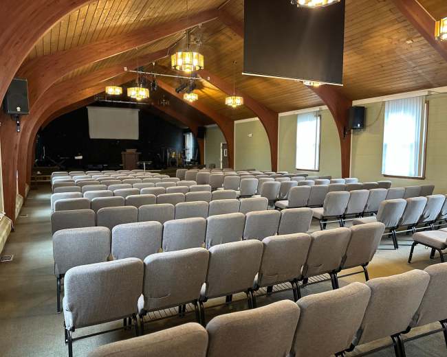 The image shows a spacious auditorium with rows of gray chairs facing a stage under wooden beams and pendant lights.