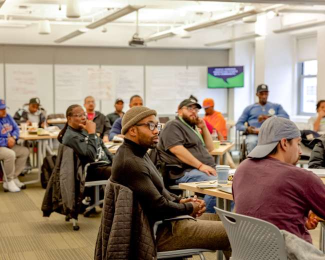 A diverse group of people is seated in a classroom setting, engaged in a discussion, with whiteboards and a presentation screen visible in the background.