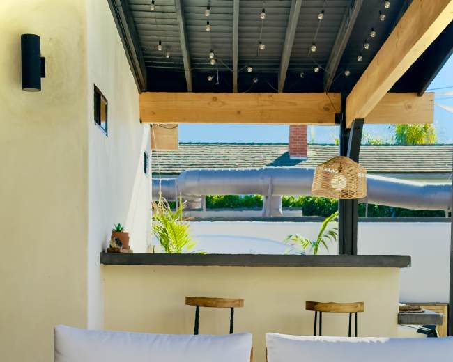 The image shows a cozy outdoor seating area featuring two white cushioned chairs facing a round fire pit, with a bar area and greenery in the background.