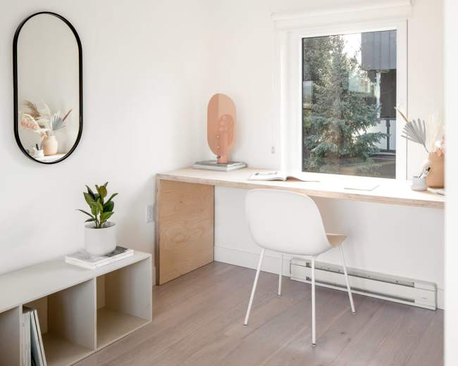 A minimalist home office features a wooden desk by a window, a chair, a mirror, a plant, and organized shelves.