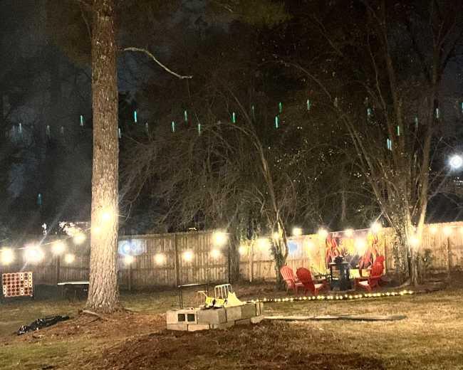 The image shows a backyard at night with string lights illuminating a seating area, surrounded by trees and grass.