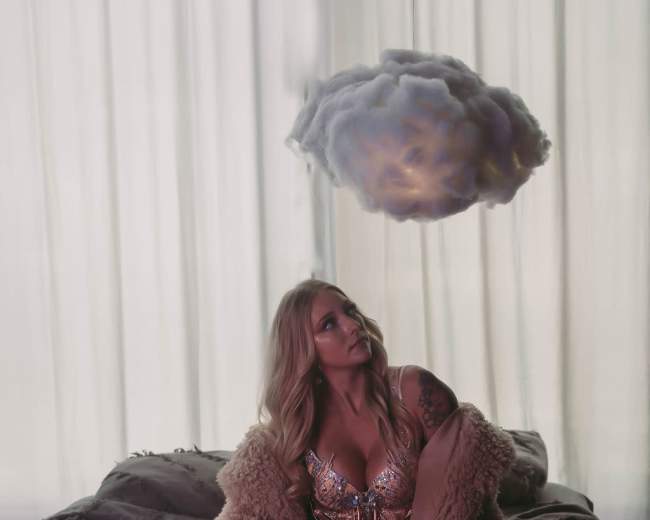 A woman in a shimmering outfit sits on a bed, gazing upward at a cloud hovering above her.