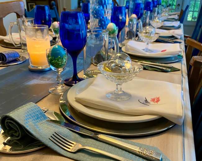 A long dining table is beautifully set with blue glassware, elegant plates, and decorative napkins in preparation for a meal.