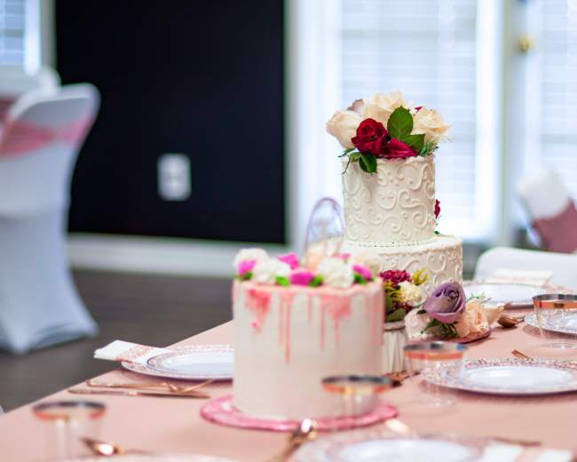 Two elegantly designed wedding cakes with decorative flowers sit on a table set with fine dishware and a pink tablecloth.
