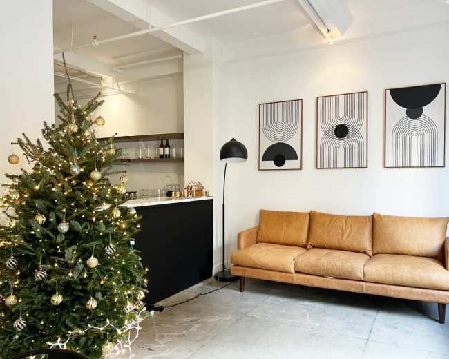 A decorated Christmas tree stands next to a brown leather sofa and three abstract framed artworks on a white wall in a modern living space.