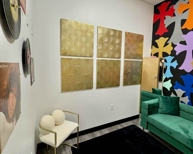 The image shows a modern lounge area with a green couch, a black rug, and decorative gold wall art, alongside a vinyl record and a colorful mural on the wall.