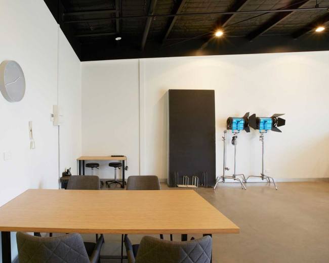 The image shows a bright, minimalist room with a wooden table and chairs, a clock on the wall, and professional lighting equipment in the corner.