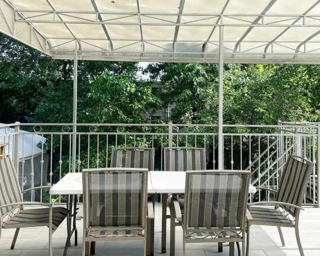 A patio with a rectangular table surrounded by six striped chairs is covered by a transparent canopy and overlooks greenery.