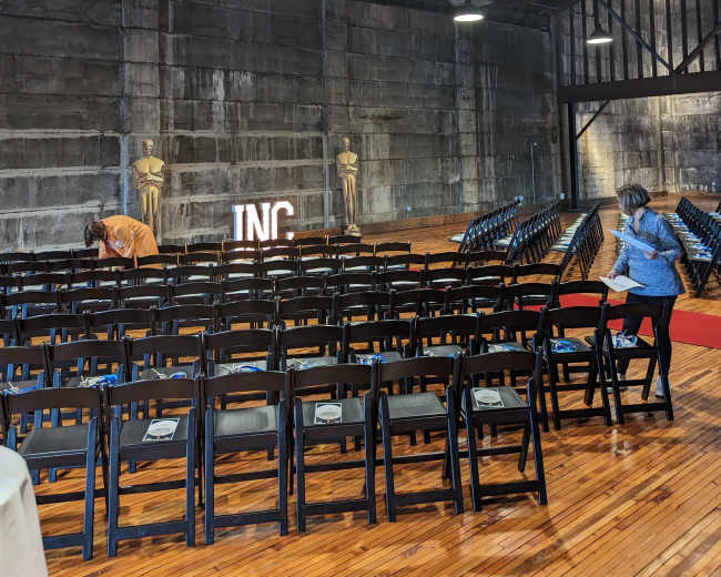 An indoor event space with rows of black chairs set up facing a lit sign that says "INC," and a few people preparing the area.