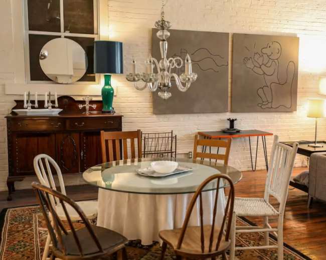 A dining area features a round table with a white tablecloth surrounded by various wooden chairs, while a chandelier hangs overhead and artwork adorns the exposed brick wall.