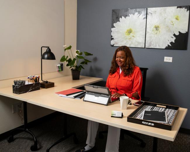 A woman in an orange blouse sits at a desk working on a tablet in an office with a flower-themed wall decoration.