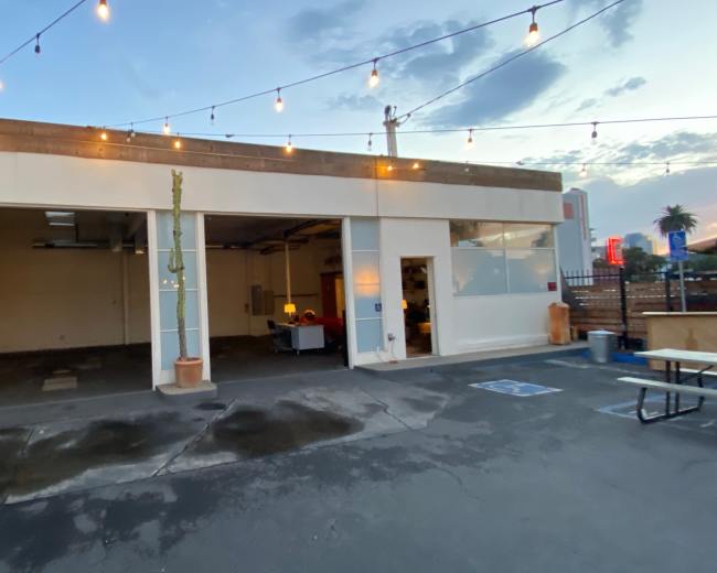The image shows a partially enclosed industrial space with large windows, surrounded by string lights and featuring a few outdoor seating arrangements.