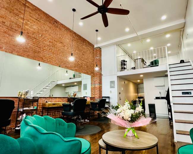 The image shows a stylish interior featuring a brick wall, green seating, a staircase leading to an upper level, and contemporary decor.